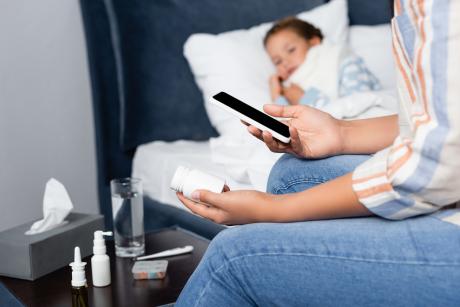 Mother holding bottle of pills and mobile phone near sick daughter lying in bed