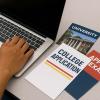 A close-up of a student’s hands typing on a laptop with college brochures nearby. 